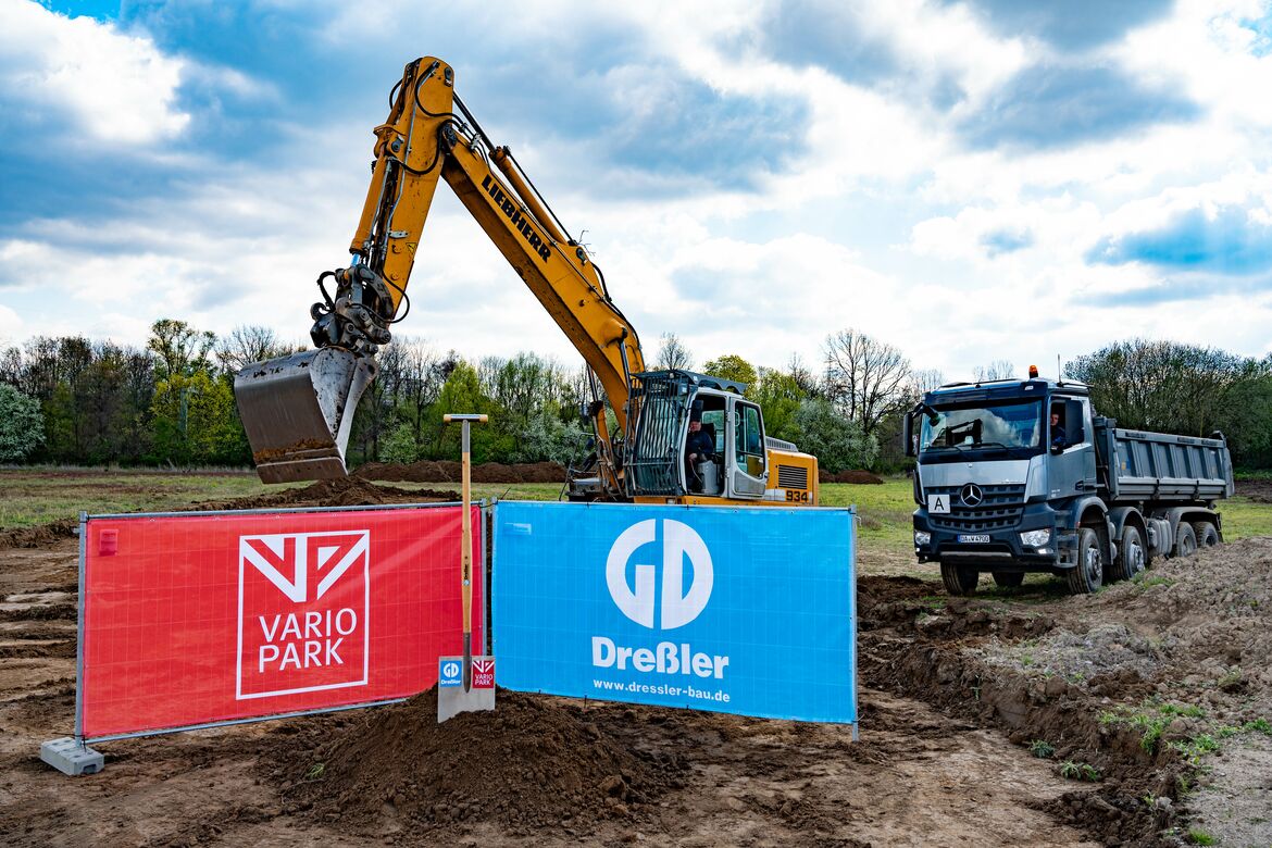 Ein riesiger Spaten mit den Logos der VarioPark Gmbh Und der Dreßler Bau GmbH in einem Erdhügel auf der Baustelle mit Bagger und Laster
