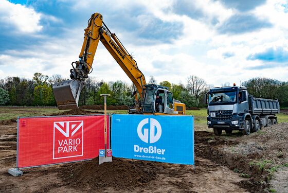 Ein riesiger Spaten mit den Logos der VarioPark Gmbh Und der Dreßler Bau GmbH in einem Erdhügel auf der Baustelle mit Bagger und Laster